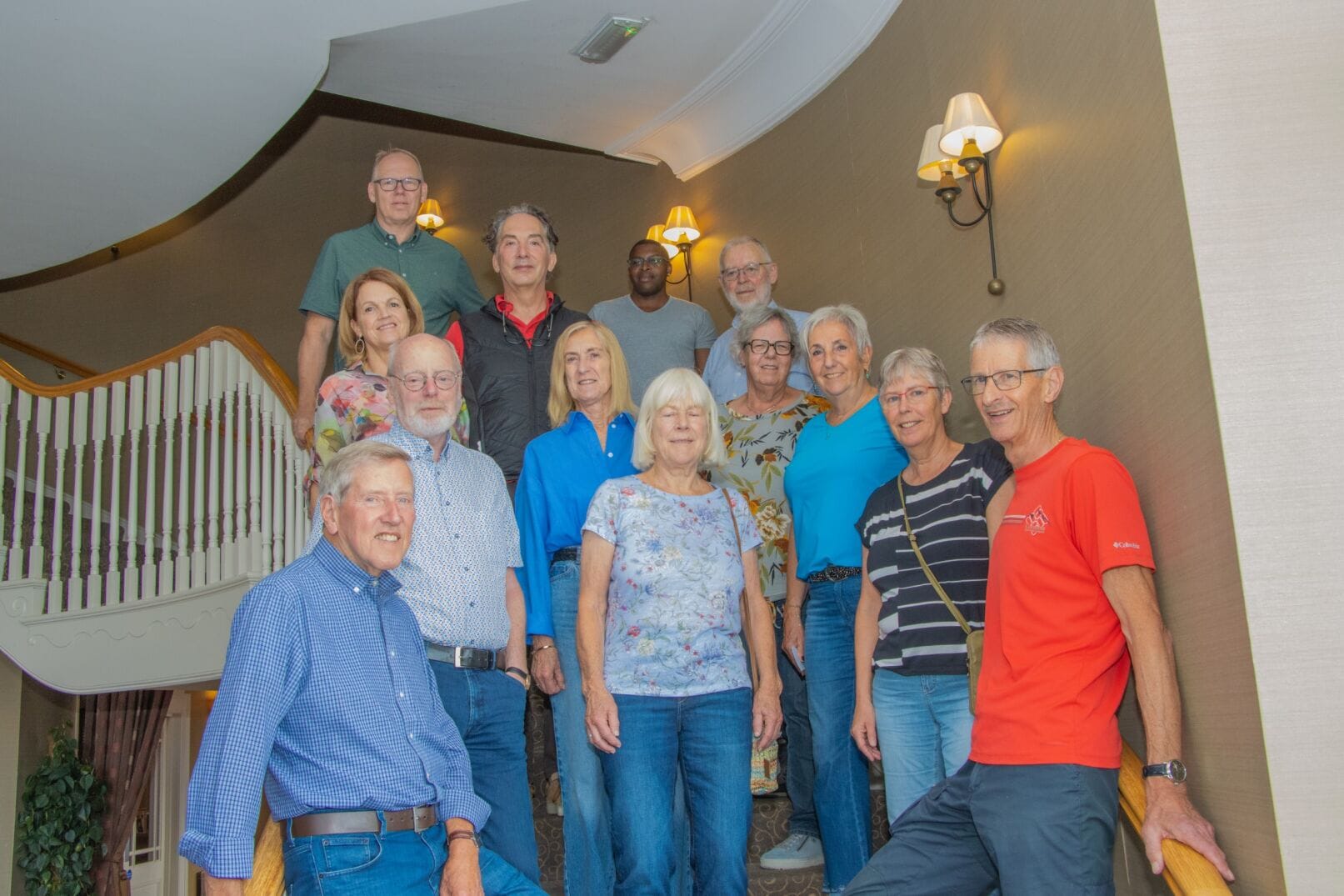 A group of older adults stands together, smiling, on a staircase inside a well-lit building with beige walls and white railing.