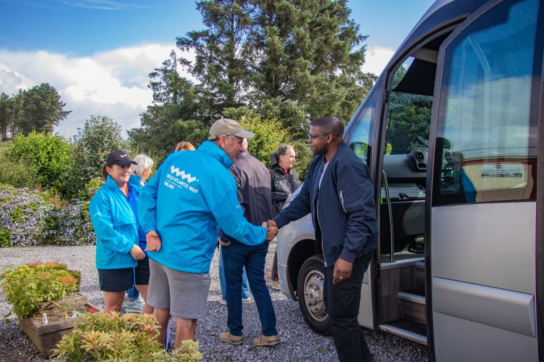 A group of people greet a man stepping out of a van on a sunny day, shaking hands and smiling outdoors.