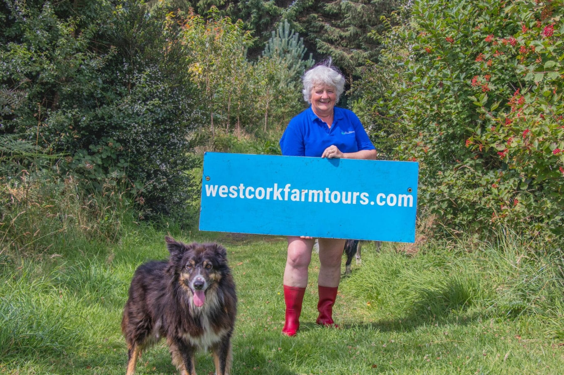 Smiling person in red boots holds a blue westcorkfarmtours.com sign; a dog stands nearby on a grassy path surrounded by trees.