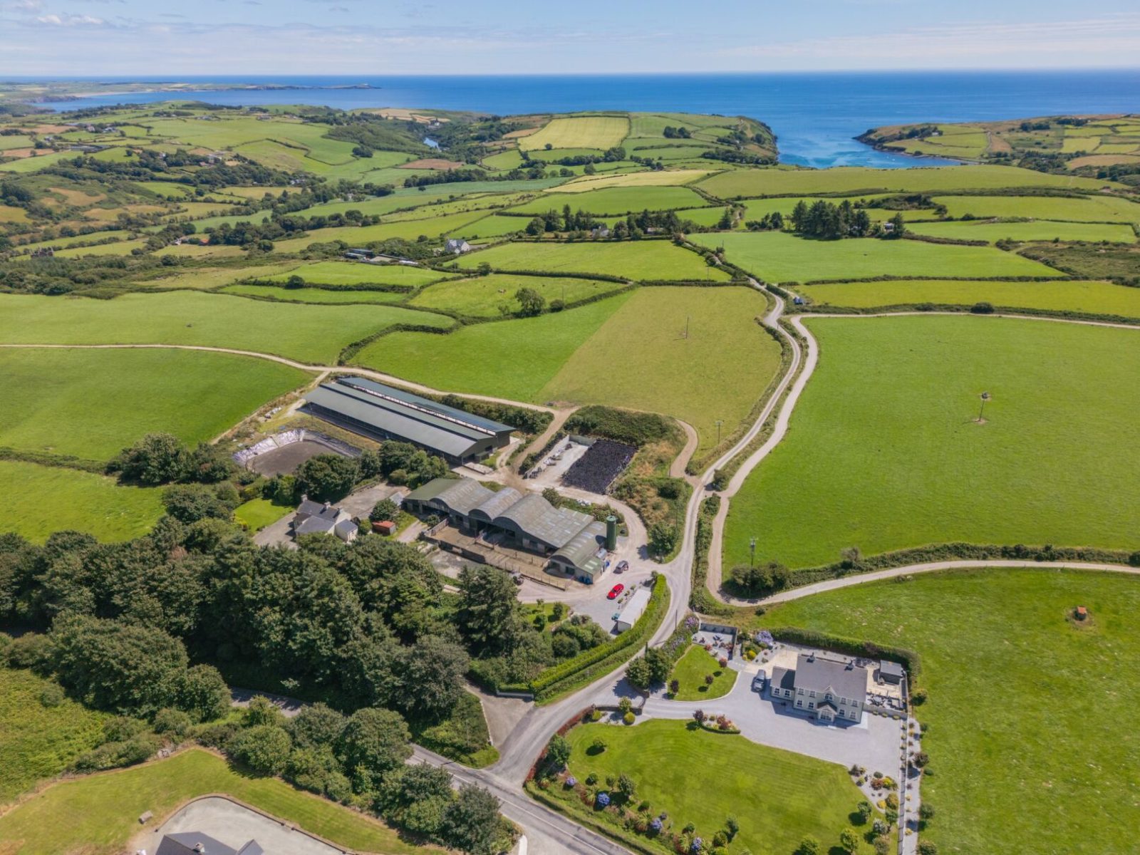 Aerial view of green fields, dairy farmers’ buildings, winding roads, and coastline under a blue sky.