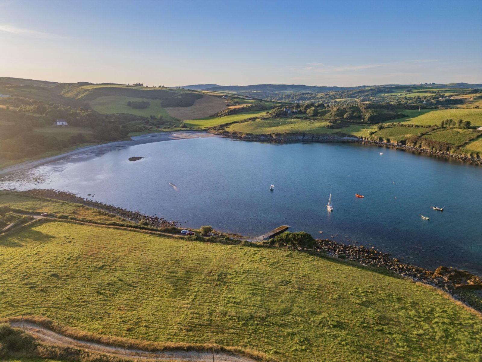 A calm coastal bay with green rolling hills, where sustainable farmers tend the land near a pebble beach and boats anchored on clear blue water.