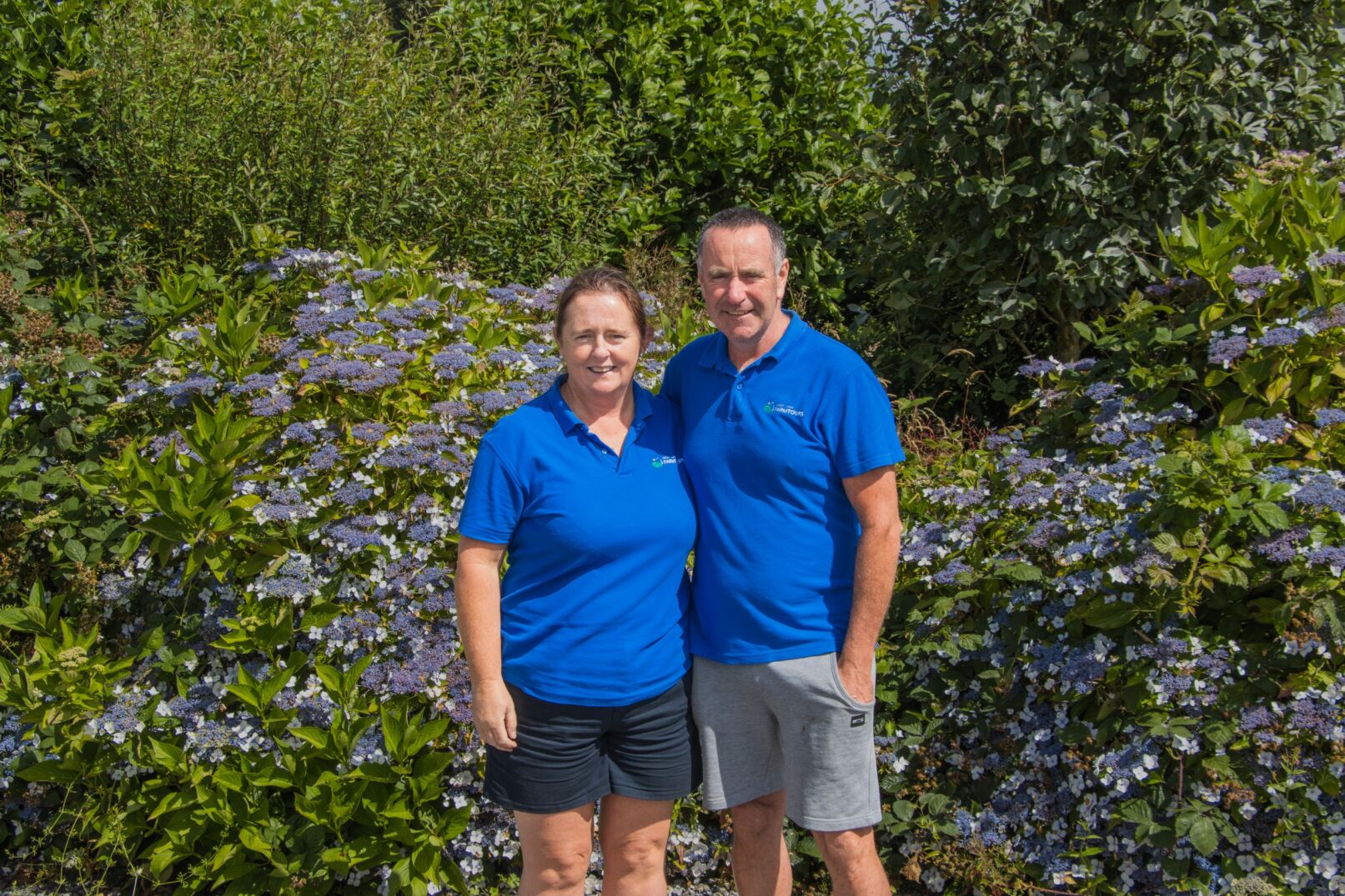 A smiling man and woman in blue shirts stand together in front of blooming bushes and greenery on a sunny day.